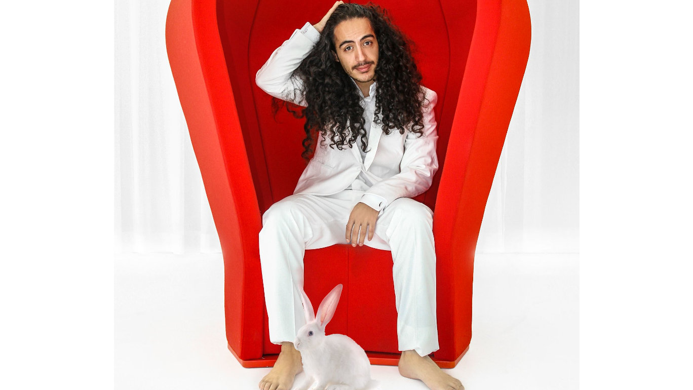 Foto von Masud im weißen Anzug, sitzend im roten Sessel, barfuß, mit weißem Kaninchen